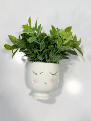 White ceramic planter with face design containing a green plant on a white surface